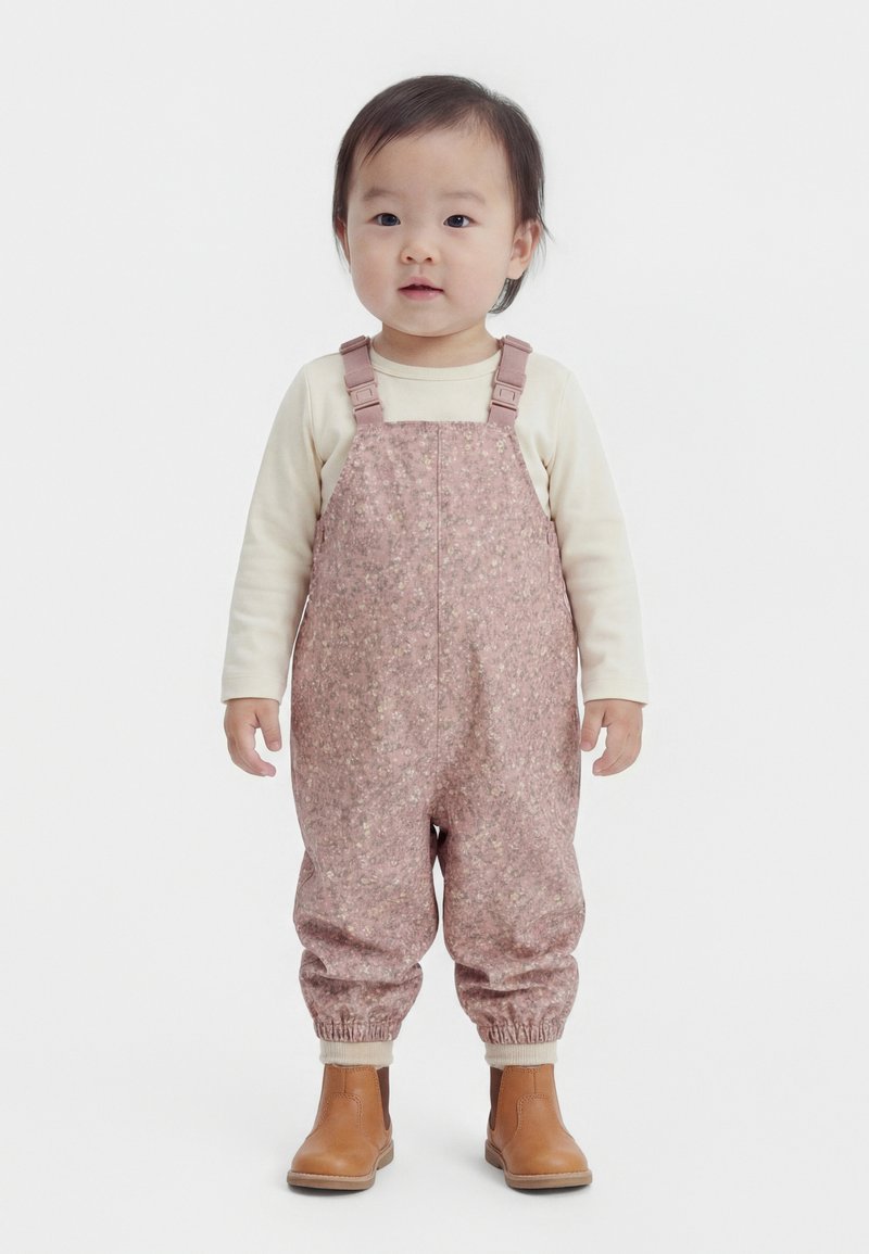 Bambino in piedi rivolto in avanti, che indossa una maglietta chiara a maniche lunghe, salopette rosa con motivi floreali e stivaletti beige alla caviglia su sfondo bianco.