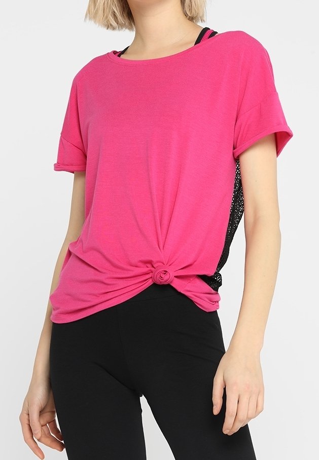 Žena v jasně růžovém krátkém rukávu tričku, uvázaném v pase, s černými síťovanými panely po stranách, v kombinaci s černými legínami.