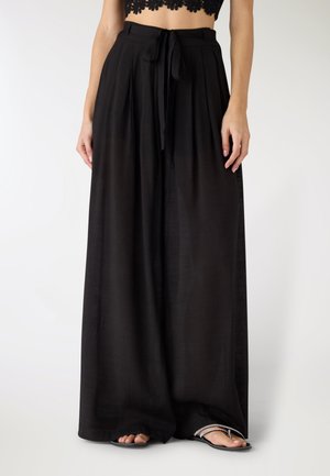 Donna che indossa pantaloni neri a gamba larga con vita alta e cintura annodata sul davanti, con sandali neri, in piedi davanti a uno sfondo neutro.