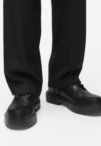 Sapatos de pele preta com bico arredondado, sola de borracha espessa e costura decorativa. Os atacadores estão segurados com ilhoses de metal.