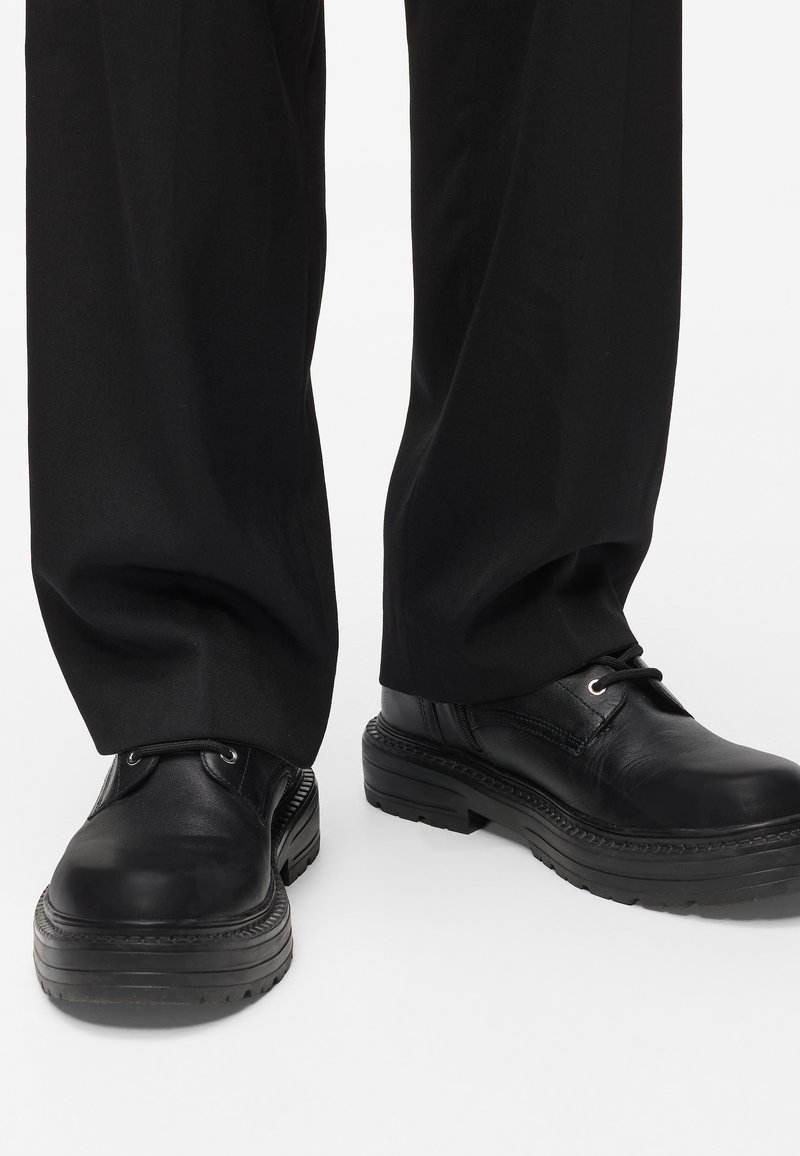 Sapatos de pele preta com bico arredondado, sola de borracha espessa e costura decorativa. Os atacadores estão segurados com ilhoses de metal.