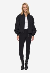 Černá bomber bunda s nafouklými rukávy a zapínáním na zip, ve spojení s černými přiléhavými kalhotami a kotníkovými botami. Hladká textura látky.