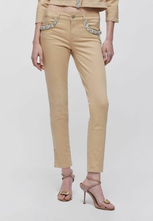 Pantaloni beige slim fit con tasche anteriori impreziosite da cristalli, indossati con giacca corta abbinata e sandali nude con tacco alto e cinturini alla caviglia.