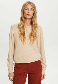 Suéter de punto beige claro con mangas largas y ligeramente abullonadas, y un escote redondeado. Combinado con pantalones de color óxido. Textura suave.