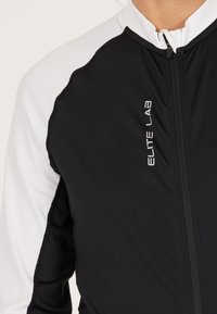 Schwarze und weiße Zip-Jacke aus strukturiertem schwarzem Stoff mit glatten weißen Schultern. Mit "ELITE LAB" Branding auf der Vorderseite.