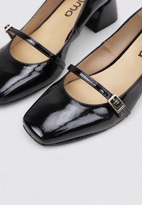 Zapatos de charol negro con tacón cuadrado, presentan una delgada correa y un detalle de hebilla dorada en la parte frontal.