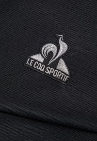Tessuto nero con una finitura texture caratterizzato da un logo ricamato in argento che mostra "Le Coq Sportif" e un design stilizzato di un gallo.