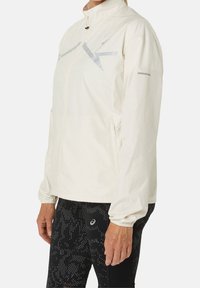 Vrouw in een crèmekleurige ritsjack met reflecterende strepen en zwarte leggings met een patroon, met ontspannen armen.