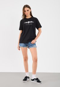Tricou negru cu mâneci scurte și un imprimeu grafic cu luni și textul "E DOAR O FAZA." Asortat cu pantaloni scurți din blugi uzati și adidași.