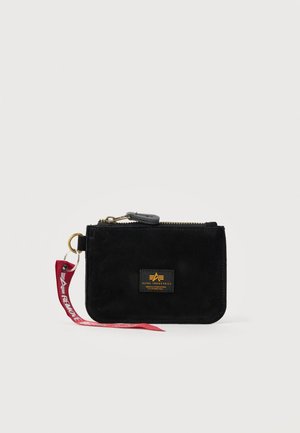 Sort lynlåslommebog med Alpha Industries logo-patch og rød stofnøglering fastgjort til guldring på ensfarvet baggrund.