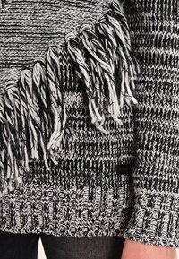 Czarny i biały sweter robiony na drutach z grubymi frędzlami po bokach, noszony na ciemnych spodniach.