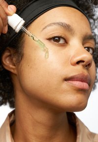 Szklana butelka z zakraplaczem wypełniona zielonym serum, aplikowana na skórę policzka modelki. Widać kręcone włosy, jasnobrązową skórę i delikatny pierścionek w nosie.