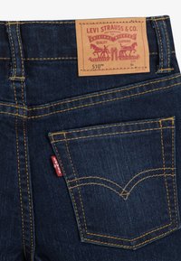 Indigo džinsa biksēm ir dzelteni šuvumi, aizmugurējā kabata ar loka dizainu un brūna ādas etiķete ar sarkanu logotipu, kas norāda izmēru 6A.