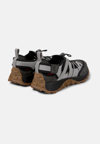 Scarpe sportive nere e grigie con una suola robusta e testurizzata, lacci elastici e tallone rinforzato, caratterizzate da un design di cinture intrecciate di forte impatto.