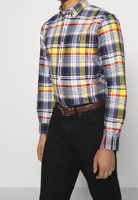 Krageknäppt skjorta med ett flerfärgat rutmönster i gult, rött, blått och grönt. Bärs med svarta byxor och ett brunt bälte.