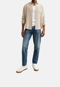 Vaaleabeige nappipaita valkoisen toppauksen päällä, yhdistetty haalistuneisiin sinisiin farkkuihin ja valkoisiin lenkkareihin, joissa on ruskeat pohjat.