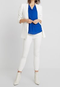 Biały żakiet na niebieskiej bluzce, w połączeniu z białymi skinny jeansami oraz białymi botkami za kostkę. Gładkie tekstury i dopasowana sylwetka.