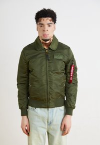 Olivno zelena bomber jakna z zadrgo spredaj, velikim ovratnikom in rebrastimi manšetami. Ima našitek in rdečo etiketo na rokavu. Prijeten, sproščen kroj.