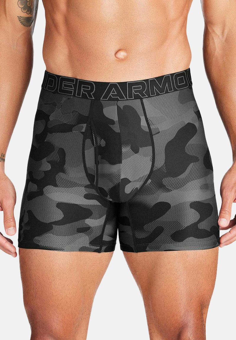 Torso masculino con bóxers Under Armour de camuflaje negro y gris, con cintura elástica con logo y tatuaje visible en el brazo izquierdo.