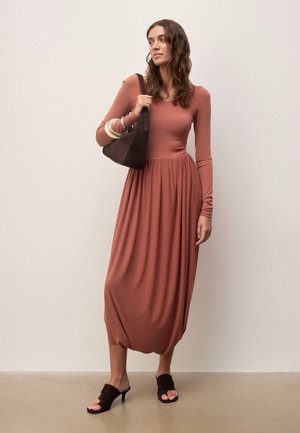 Femme portant une robe midi à manches longues de couleur rouille, tenant un sac à épaule noir, chaussée de sandales à talons noires, debout devant un mur uni.