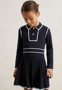 Marineblaues Kleid mit langen Ärmeln, weißen Akzenten, geflochtenen Details am Kragen, einem plissierten Rock und Knopfverschluss.