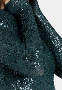 Teal paljettopp med en texturerad stickad design, långa ärmar och en figurnära silhuett med skimrande metalliska accenter.