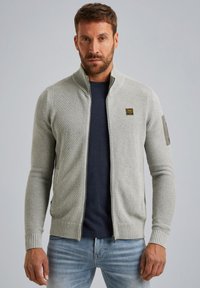 Lichtgrijze ritsvest met ribbeltextuur, hoge kraag en subtiele logo-detail op de arm; gecombineerd met een marineblauw shirt en blauwe spijkerbroek.