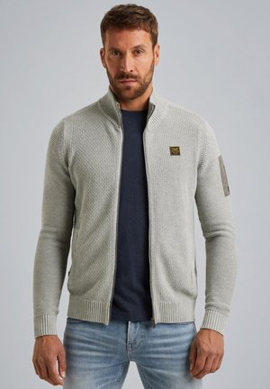 Hellgraue Zip-Up-Strickjacke mit gerippter Textur, hohem Kragen und dezentem Logodetail am Arm; kombiniert mit einem navyblauen Shirt und blauen Jeans.