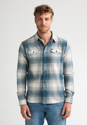 Mann mit lockigem Haar und Bart, der ein blau-weiß kariertes Hemd mit Knöpfen und blaue Jeans trägt, steht vor einem einfarbigen Hintergrund.