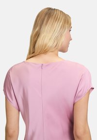 Helles rosa Kleid mit einem weichen Stoff, kurzen Ärmelchen und einem verdeckten Reißverschluss auf der Rückseite. Glatte Textur mit schlichten Designelementen.