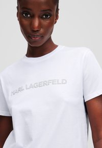 Weißes Baumwoll-T-Shirt mit metallic-silbernem "KARL LAGERFELD"-Text auf der Vorderseite, Rundhalsausschnitt und kurzen Ärmeln. Glatte Textur.