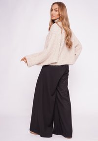 Kobieta z długimi blond włosami, ubrana w beżowy, oversize'owy sweter i czarne, szerokie spodnie, stoi na tle jednolitego, jasnego tła.