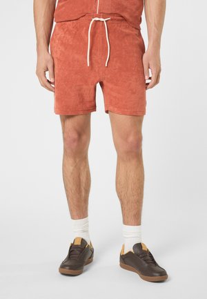 Uomo che indossa pantaloncini in spugna color ruggine con coulisse bianche, calzini bianchi e scarpe da ginnastica marroni su uno sfondo chiaro uniforme.