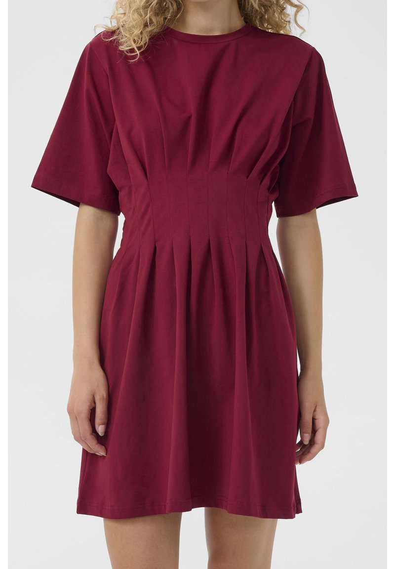 Femme portant une robe rouge foncé à manches courtes, longueur genou, avec des plis à la taille et un col rond sur un fond uni.