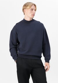 Tengerészkék pulóver magas gallérral, laza szabással és bordázott mandzsettával. Puha szövettextúra, minimalista design, nincs látható minta vagy grafika.