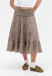 Brązowa plisowana spódnica warstwowa z elastycznym paskiem w talii, z falistym wykończeniem na dole. Noszona z jasnymi skarpetkami i zielonymi butami.