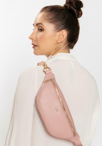 Hellbraune Leder-Sling-Tasche mit goldfarbenem Reißverschluss, mit strukturiertem Finish und verstellbarem Riemen, getragen über einem weißen, gerippten Oberteil.