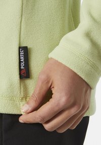 Jasnozielony polarowy top o teksturowanej powierzchni, z przeszywanymi szwami i widoczną czarną metką "POLARTEC" blisko dolnego brzegu. Ręka dostosowuje materiał.