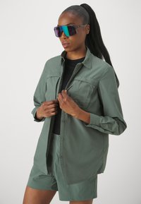 Camicia verde chiaro con bottoni e due tasche sul petto, indossata sopra a una maglietta nera e pantaloni corti abbinati. Grande occhiali da sole riflettenti con gradiente.