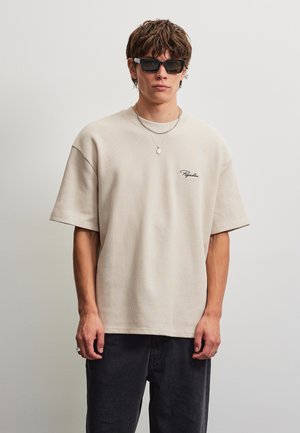 Junger Mann mit schwarzen Sonnenbrillen, einem beigefarbenen gerippten T-Shirt mit kleinem Logo, dunkler Hose und geschichteten silbernen Halsketten, der vor einer schlichten Wand steht.