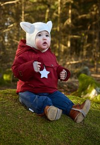 Chaqueta impermeable roja con dos bolsillos frontales, gorro blanco, pantalones azules y botas marrones, sentado en el suelo cubierto de musgo con árboles de fondo.