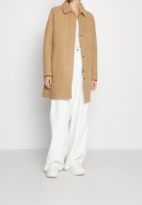 Kamelfarbener Mantel aus weichem Material mit Kragen, Druckknöpfen und seitlichen Taschen. Kombiniert mit weißen weiten Hosen und Turnschuhen.