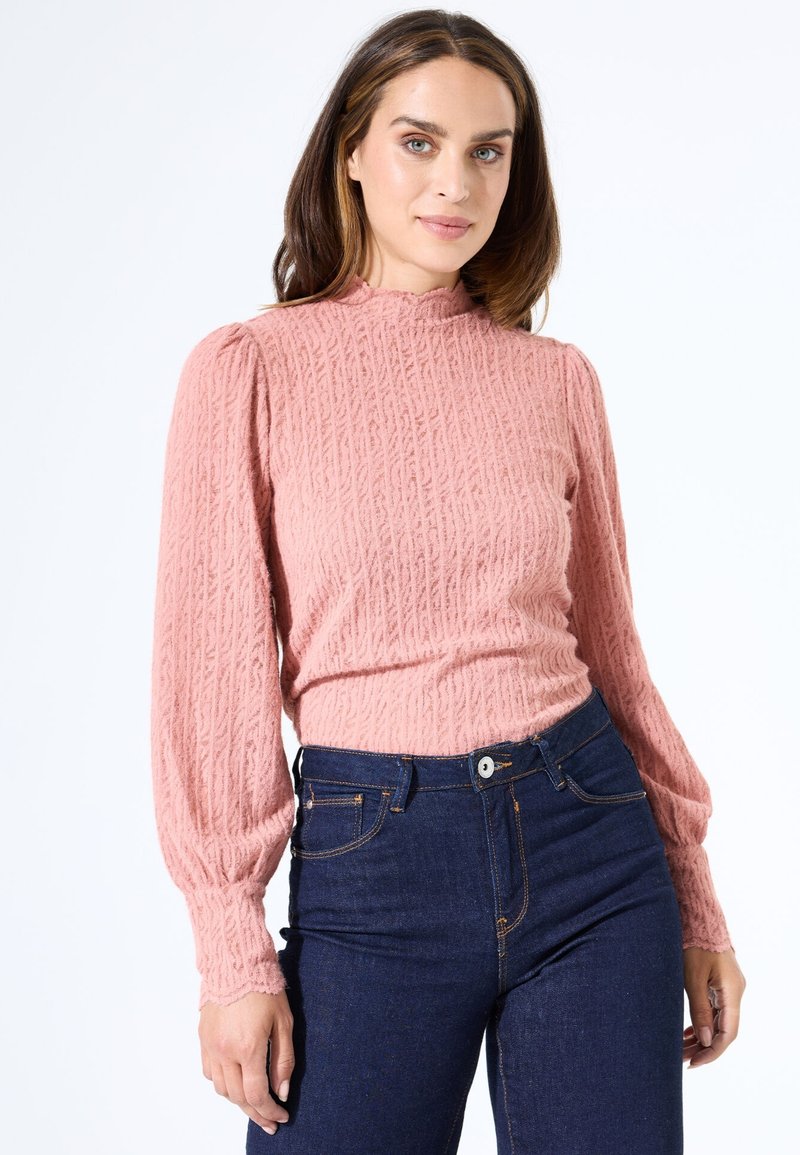 Rosa strukturiertes Langarmoberteil mit hohem Kragen und gepufften Schultern, kombiniert mit dunkelblauen, figurbetonten Jeans. Mit gewellten Bündchen.