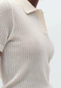 Strick-Polohemd mit kurzen Ärmeln in hellbeige, mit geripptem Kragen und Knopfdetails. Der Stoff hat eine offene, atmungsaktive Textur.