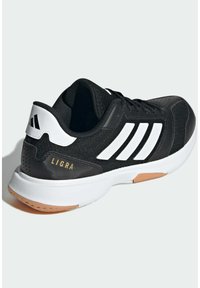 Chaussure de sport Adidas Ligra noire et blanche avec semelle blanche et semelle extérieure couleur gomme, vue sous un angle arrière latéral sur un fond uni.