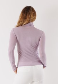 Top a dolcevita lilla realizzato in tessuto morbido e elasticizzato. Maniche lunghe e design aderente. Indossato con pantaloni bianchi, visto da dietro.