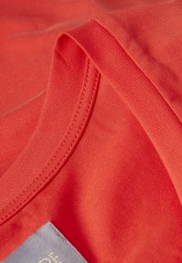 Red cotton fabric with a soft texture, featuring a ribbed crew neckline and a gray label at the bottom left corner.