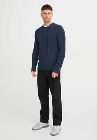 Pull en maille bleu marine avec un motif de cable texturé, col rond et poignets côtelés, associé à un pantalon noir et des baskets grises.