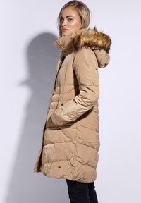 Dlouhý krémový péřový kabát se zipem a patentkami, s odnímatelnou kapucí podšitou kožešinou a bočními kapsami. Strukturovaný prošívaný design.