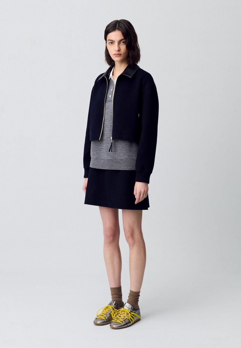Chaqueta corta azul marino con cremallera dorada, camisa gris en capas debajo, falda oscura y zapatillas metálicas con cordones amarillos.
